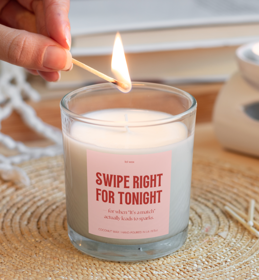 Hand lighting a 'Swipe Right for Tonight' candle by lol wax, with a playful pink label, hand-poured coconut wax, and cozy wicker background.