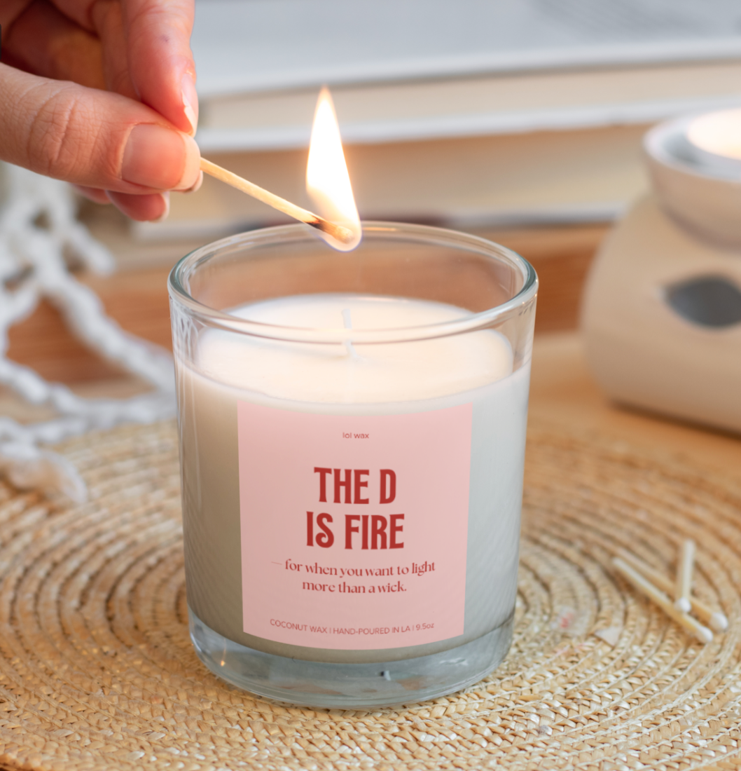 lol wax's 'The D is Fire' candle with a sassy pink label, a flame softly glowing, setting a flirty mood.