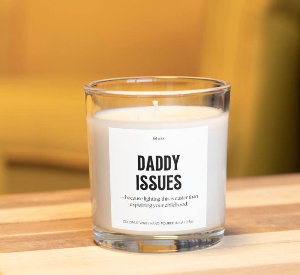 A 'Daddy Issues' candle by lol wax held in hand against a mustard yellow background, highlighting the playful quote on the label.