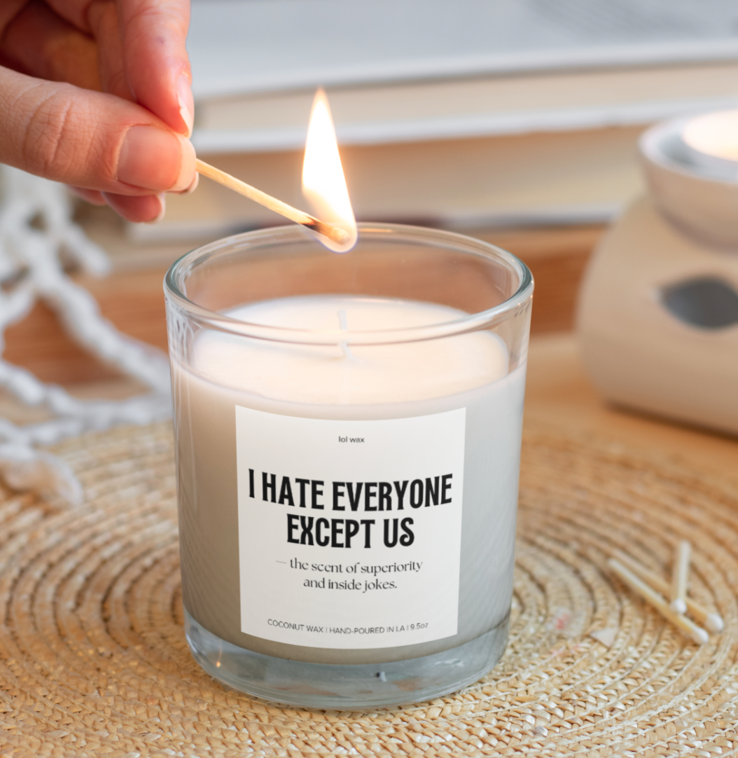 A hand lighting the 'I Hate Everyone Except Us' candle by lol wax, placed on a woven mat, evoking a cozy, intimate vibe.