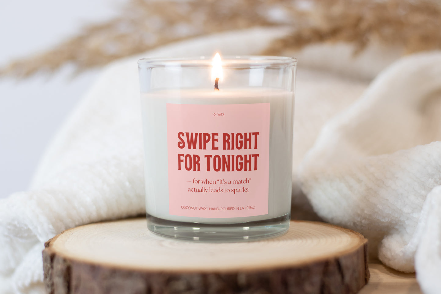 A serene setting featuring 'Swipe Right for Tonight' candle by lol wax on a wood slice, surrounded by a soft white blanket, inviting a warm, relaxing experience.