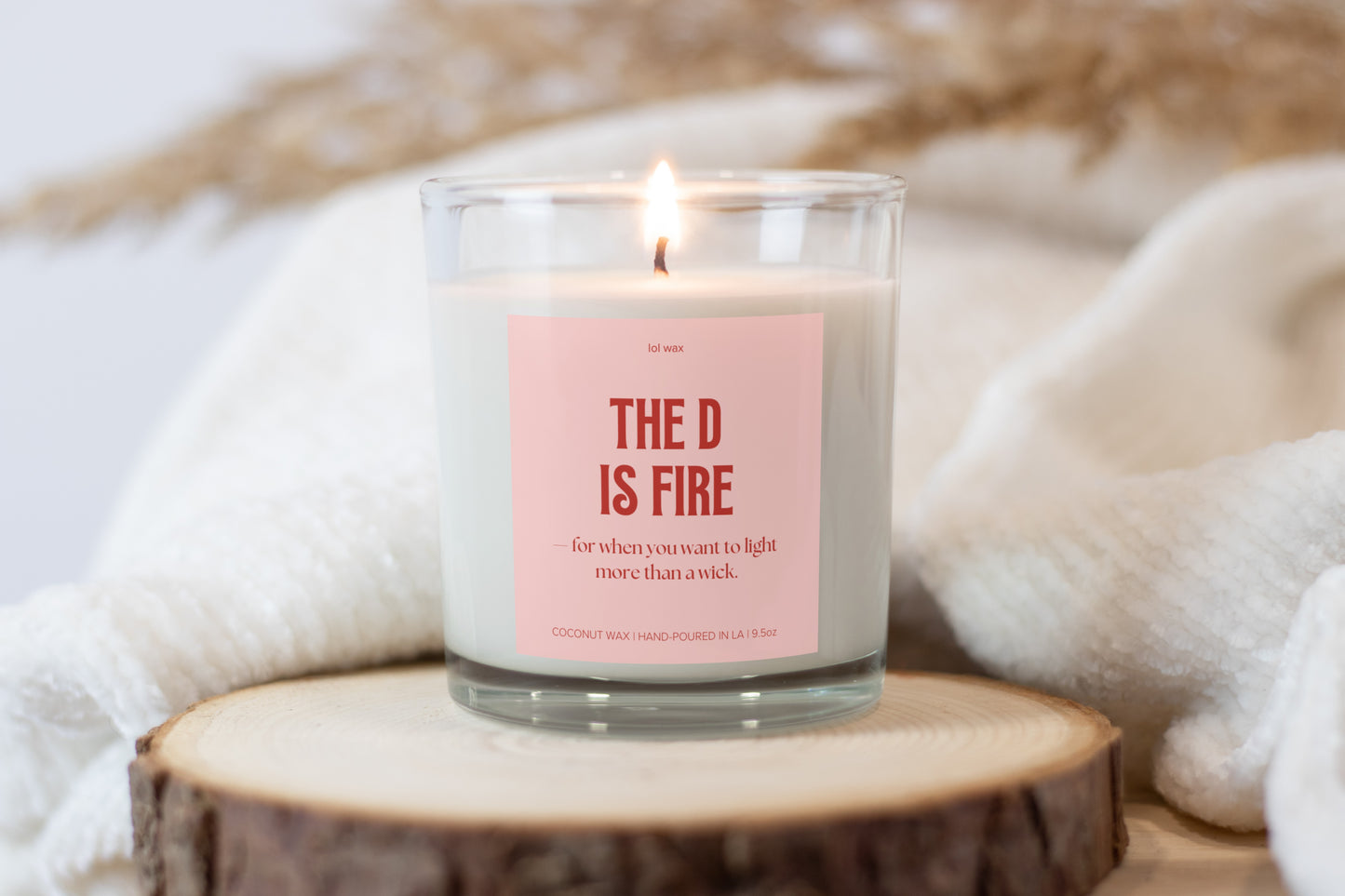 Cozy ambiance with 'The D is Fire' candle by lol wax on a wooden coaster, complemented by a plush white blanket and a rustic backdrop.