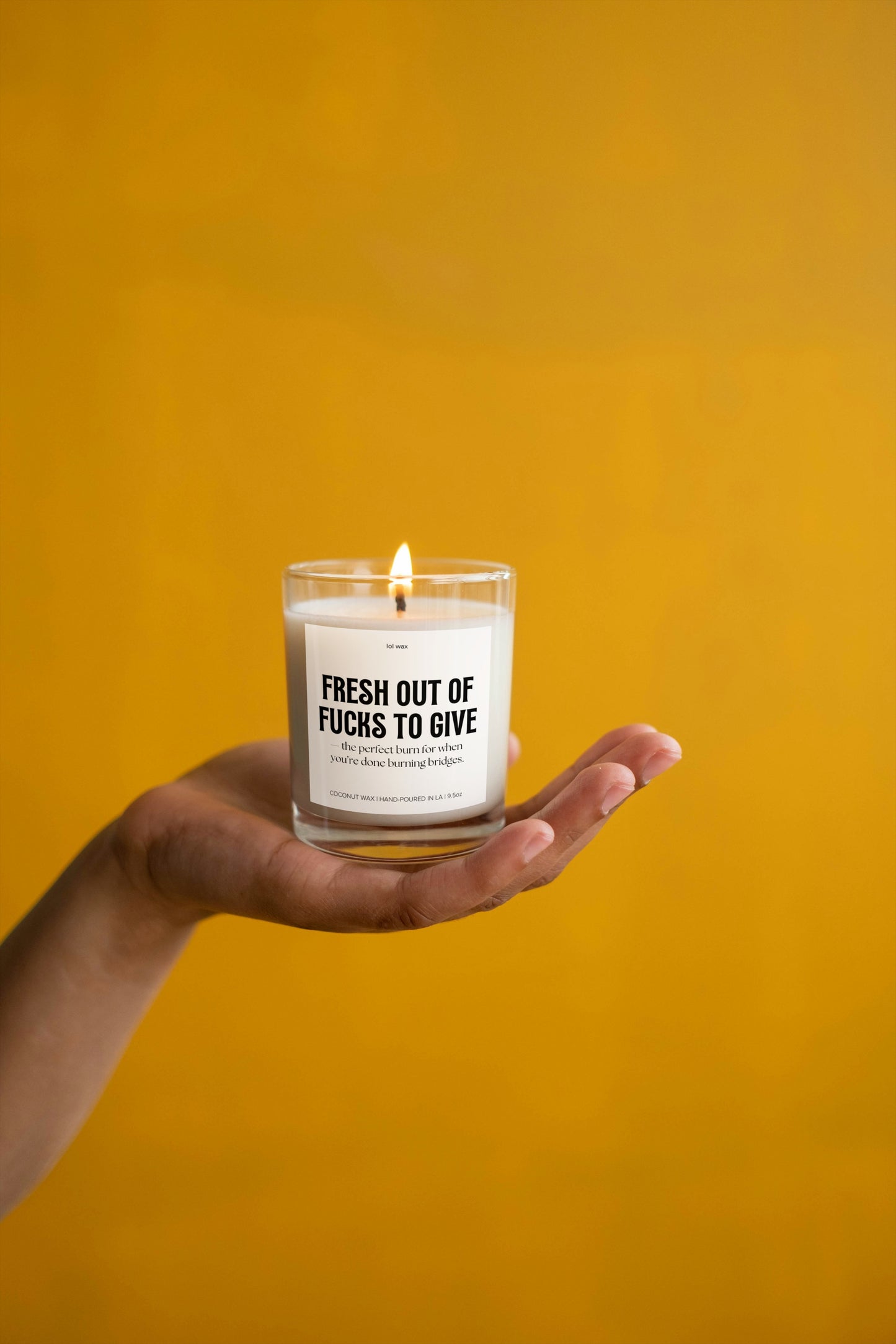 A hand holding a 'Fresh Out of Fucks to Give' candle by lol wax against a bright yellow background, symbolizing warmth and energy.